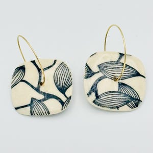 May include: A pair of square earrings with a cream-colored base and a dark blue leaf pattern. The earrings have gold-colored hoops. The leaf design is a repeating pattern of stylized leaves and branches.