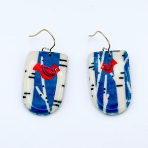 May include: A pair of handcrafted earrings featuring a red cardinal bird design. The earrings have a rectangular shape with a blue and white birch tree pattern. The earrings have gold-colored ear wires.