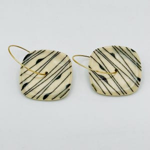 May include: A pair of square earrings with a cream-colored base and black diagonal lines. The earrings have gold-colored hoops. The design features small black accents along the lines, adding a unique touch to the jewelry.