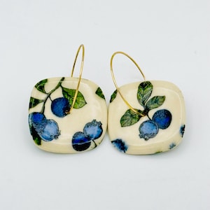May include: A pair of square earrings with a cream-colored background. Each earring features a design of blue blueberries and green leaves. The earrings have gold-colored hoop closures.