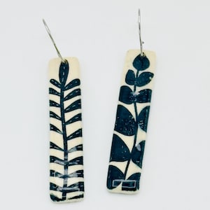 May include: A pair of rectangular earrings with a cream-colored base and a dark blue leaf design. The earrings have a silver-colored hook. The leaf design is a stylized botanical pattern.