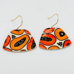 May include: A pair of earrings with a unique papaya design. The earrings feature a cream-colored base with orange and black papaya fruit illustrations. The earrings have gold-colored hooks.