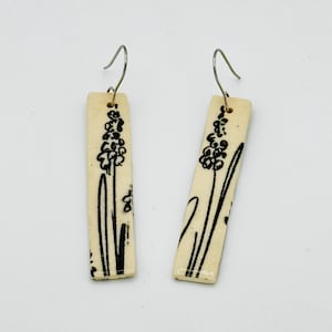 May include: A pair of rectangular earrings with a cream-colored base. Each earring features a black floral design. The earrings have silver-colored hooks. The floral design appears to be a stylized hyacinth.