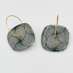 May include: A pair of square earrings with a gold hoop. The earrings have a black and white circular pattern. The earrings are on a white background.