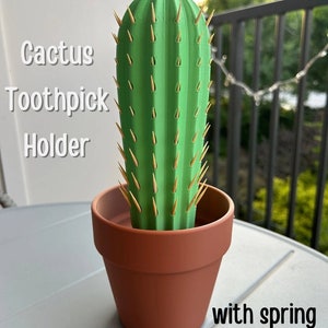 以下が含まれることがあります： 茶色の鉢に入った緑色のサボテン型のつまようじ立て。サボテンにはつまようじを保持するためのバネ機構があります。画像には「Cactus Toothpick Holder」というテキストが表示されています。