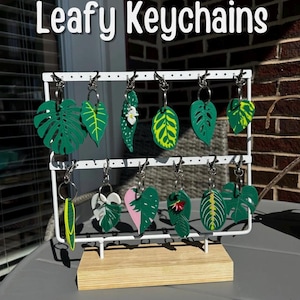 Op de afbeelding: Een display van blad-vormige sleutelhangers. De sleutelhangers zijn in verschillende groentinten, met gele, witte en roze accenten. Ze hangen aan een witte metalen displaystandaard. De tekst "Leafy Keychains" staat bovenaan.