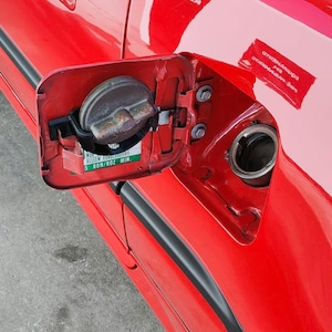 Può includere: Primo piano dello sportello del carburante aperto di un'auto rossa, che rivela il tappo del serbatoio e il bocchettone di riempimento. Il tappo è metallico e ha un adesivo verde. La carrozzeria è lucida.