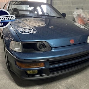 May include: A blue coupe car with the text "Magnna 30" on the hood. The car has a black front bumper, yellow fog lights, and a red emblem. The car is parked in a garage.