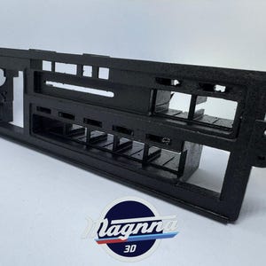 Puede incluir: Un componente negro y rectangular del salpicadero de un automóvil con múltiples aberturas y una superficie texturizada. El componente tiene una serie de ranuras rectangulares y una abertura circular en el lado izquierdo. El logotipo "Magnna 3D" es visible en la parte inferior.