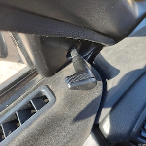 May include: Close-up of a car's interior, featuring a black lever with a textured grip. The lever is positioned near an air vent and other interior components, all in shades of black and gray. The image highlights the car's dashboard details.