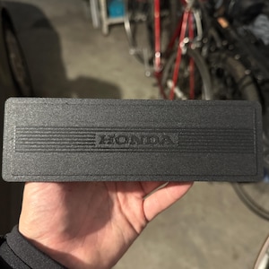 Pode incluir: Uma peça retangular preta de carro com a palavra "HONDA" em relevo no centro. A peça tem uma superfície texturizada com linhas horizontais. O fundo é uma garagem com bicicletas.