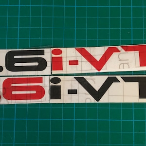 May include: Two vinyl stickers with the text "1.6 i-VT" in black and red lettering. The top sticker has red lettering and the bottom sticker has black lettering.