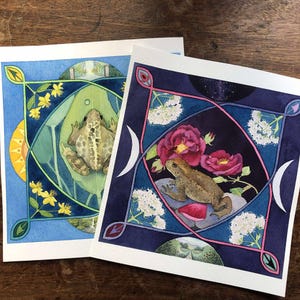 Könnte beinhalten: Zwei Aquarelldrucke mit detaillierten Illustrationen von Fröschen. Ein Druck zeigt einen Frosch auf einem Blatt mit gelben Blumen und blauem Hintergrund. Der andere zeigt einen Frosch mit rosa Rosen und einem dunkelblauen Hintergrund.