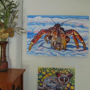 May include: A painting of a large red and yellow crab on a blue and white background. The crab is walking on a sandy beach. The painting is framed and hanging on a white wall.