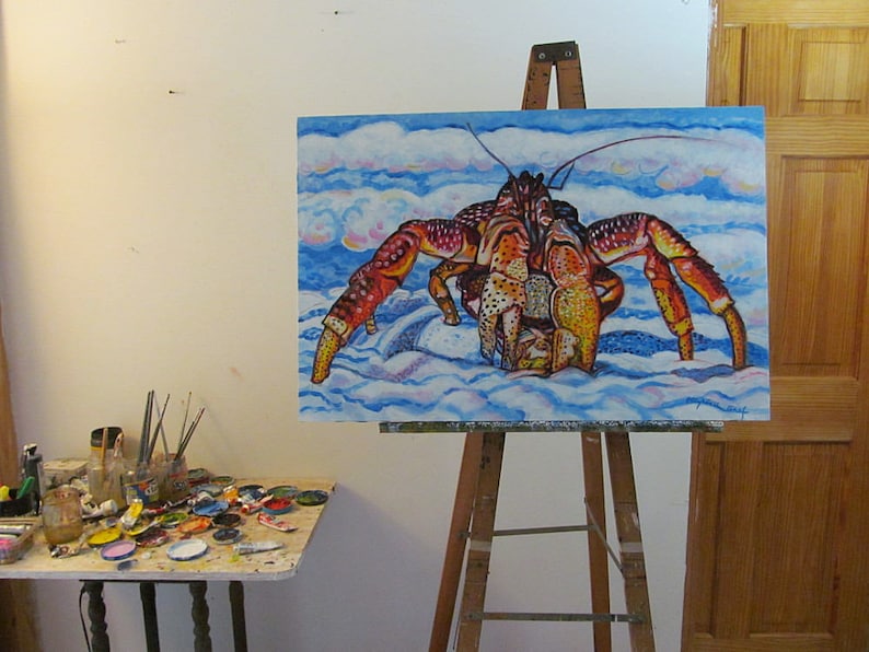 May include: A colorful painting of a large red and orange crab on a blue and white background. The crab is walking on a sandy beach with clouds in the sky. The painting is on an easel in an art studio.