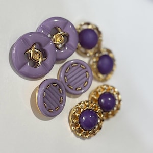 以下が含まれることがあります： さまざまなデザインと色の装飾ボタンのコレクション。ボタンには、金色のアクセントが付いた丸いラベンダー色のボタンと、紫色の中心部が付いた金色の縁取りのボタンが含まれます。ボタンは白い表面に配置されています。