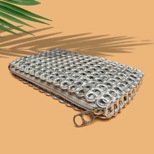 alt= silver pop tab purse with a zipper top is lying flat on an orange table a tropical green palm leaf is above the bag and casting a long shadow over the orange table