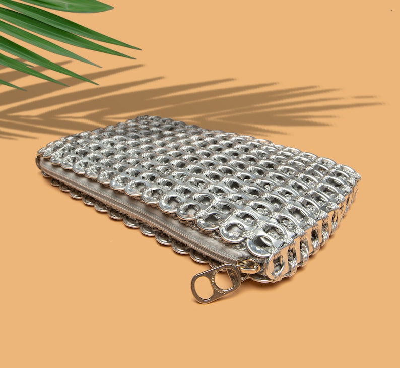 alt= silver pop tab purse with a zipper top is lying flat on an orange table a tropical green palm leaf is above the bag and casting a long shadow over the orange table