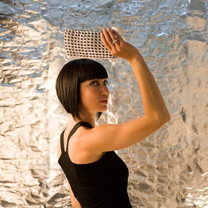 alt= a woman with a vidal sassoon bob haircut and dressed in a black evening dress is holding a metal clutch made of aluminum soda pop tabs the background is a wall made of silver aluminum