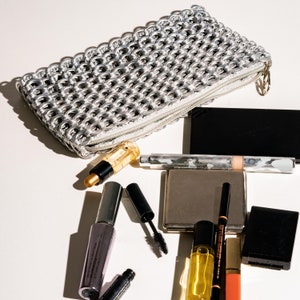 silver makeup bag made of up-cycled aluminum soda tabs is lying open on a table the contents of the clutch bag are in front and include cosmetics lip gloss perfume and a compact mirror