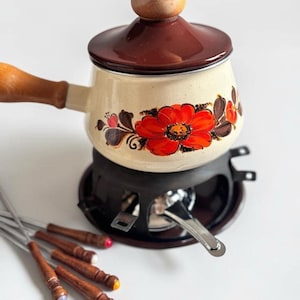 Peut inclure: Une caquelon à fondue en émail crème avec un couvercle marron et un manche en bois, ornée d'un grand motif floral rouge. Elle repose sur un support en métal noir avec un brûleur, accompagnée de quatre fourchettes à fondue en métal.