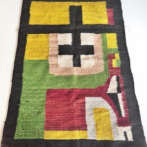 May include: A rectangular woven rug featuring a bold geometric pattern with a black border. The design includes blocks of yellow, green, red, and white, with a prominent black cross shape centered within a light brown square on a green background.