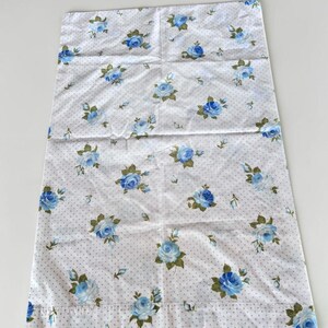 May include: A rectangular pillowcase featuring a white fabric base with a pattern of small grey polka dots and scattered blue rose blossoms with green leaves. The fabric has a smooth texture and a hemmed edge along the bottom of the rectangular shape.