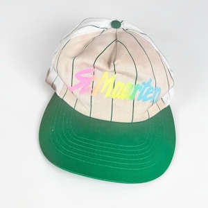 May include: A baseball cap featuring a beige crown with thin vertical green pinstripes and a solid green brim. The front of the cap displays the text St. Maarten in a multi-coloured gradient font, and a small green button sits at the top centre.