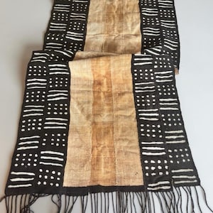 May include: A rectangular textile runner featuring a central tan panel flanked by two black side panels. The black sections are decorated with white hand-painted patterns of horizontal lines and dots. The bottom edge has long black fringe tassels.