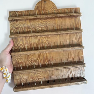 Peut inclure: Un présentoir mural rectangulaire en bois avec une finition naturelle aux tons brun clair et un grain visible. Il comporte quatre étagères horizontales, chacune équipée d'une série de tiges métalliques verticales régulièrement espacées.