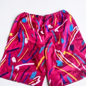 Può includere: Un paio di pantaloncini rosa acceso caratterizzati da un motivo astratto di pennellate sparse nei toni del blu, giallo, bianco e viola scuro. I pantaloncini presentano un elastico in vita e una vestibilità ampia con orlo dritto.
