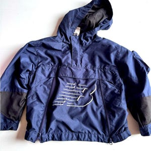 Peut inclure: Un coupe-vent à enfiler bleu marine doté d'une capuche, de manches longues avec des empiècements en maille noire aux coudes et d'un grand logo New Balance blanc brodé sur la poche centrale avant. La veste inclut une fermeture éclair partielle.