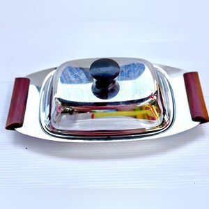 Peut inclure: Un beurrier rectangulaire en acier inoxydable avec une finition polie réfléchissante. Le couvercle comporte un bouton noir central, et la base comprend deux poignées latérales en bois brun foncé fixées aux extrémités du plateau métallique.