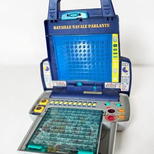 May include: A portable electronic Battleship game with a dark blue folding case and a light grey base. The unit features a vertical grid screen, a horizontal playing board with clear plastic cover, and control buttons labelled BATAILLE NAVALE PARLANTE.