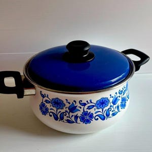 Peut inclure: Une marmite en métal émaillé blanc avec un motif floral bleu autour de la base. La marmite comprend un couvercle bleu assorti avec un bouton noir et deux poignées latérales noires. Le motif se compose de fleurs et de feuilles bleues.
