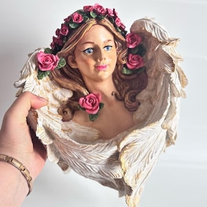 Peut inclure: Un pot de fleurs décoratif en forme d'ange avec un visage de femme, orné d'une couronne florale et de roses roses. Les ailes sont détaillées avec des textures de plumes et des accents dorés. Le pot est tenu par une main.
