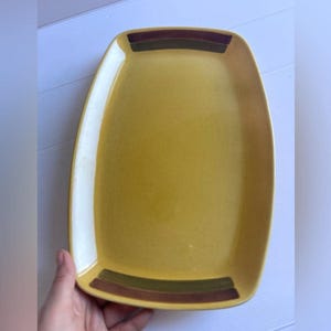 Peut inclure: Un plat de service rectangulaire en céramique avec un centre jaune moutarde et de larges bandes horizontales marron foncé et vert olive sur les bords supérieur et inférieur. Le plat présente des coins arrondis et une finition lisse et brillante.