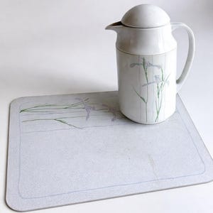 Peut inclure: Une carafe en céramique blanche avec un couvercle bombé et une anse, ornée d'un motif floral délicat d'iris violet clair et de tiges vertes. La carafe est posée sur un set de table rectangulaire gris clair avec un motif floral assorti.