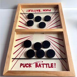Può includere: Un tabellone da gioco in legno con due sezioni separate da un divisorio centrale. Ogni lato ha una superficie chiara con linee rosse a raggiera e la scritta PUCK BATTLE! in rosso. Dieci dischi neri sono disposti sul tabellone.