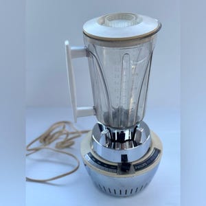 Peut inclure: Un mixeur vintage Philips avec une base chromée, un pichet en plastique transparent avec des graduations en millilitres et litres, une poignée en plastique blanc, un couvercle blanc et un cordon d'alimentation beige avec une prise électrique.