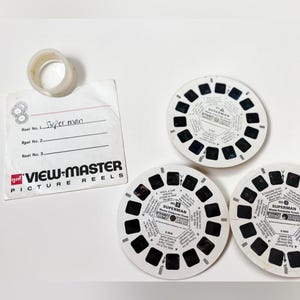 Peut inclure: Ensemble de bobines de photos View-Master vintage sur le thème de Superman. Les bobines sont blanches avec des fenêtres carrées noires et du texte. Une étiquette avec le texte "Superman" et une bague en plastique transparent sont également présentes.