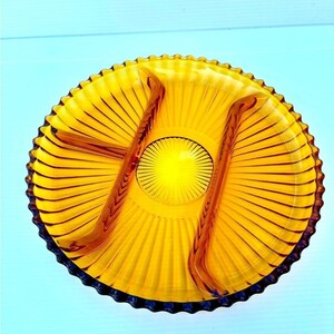 Peut inclure: Un plat de service rond en verre ambré doté d'un bord festonné et d'un intérieur divisé. La surface est ornée d'un motif rayonnant en forme d'étoile qui s'étend d'un médaillon circulaire central vers le bord extérieur de l'assiette.
