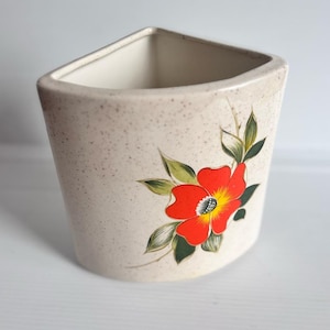 Peut inclure: Un pot de fleurs en céramique beige avec une finition mouchetée et un dessus triangulaire. Le pot présente une fleur rouge vif avec des détails jaunes et noirs, entourée de feuilles vertes. Une pièce décorative pour plantes d'intérieur.