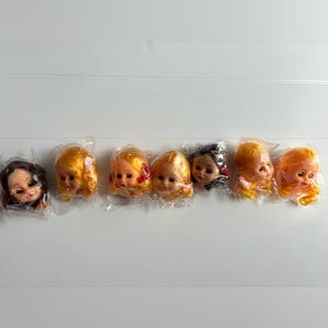 May include: A selection of vintage doll heads, individually wrapped in clear plastic. The heads display different hair colours, including blonde, brown, and black. Some have red bows. The dolls have painted facial features.