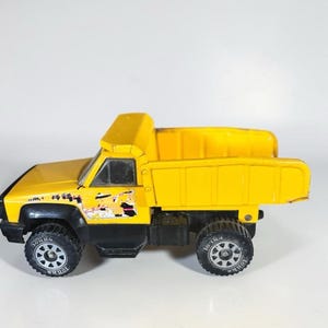 Peut inclure: Un camion benne jouet Tonka jaune avec des détails noirs. Le camion a quatre pneus noirs avec des jantes argentées et le mot "Tonka" imprimé sur les pneus. La benne est relevée. Un petit graphisme est visible sur le côté.