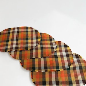 Peut inclure: Quatre dessous de verre ovales en tissu avec un motif à carreaux dans des tons orange, jaune, marron, noir et blanc. Chaque dessous de verre présente une finition effilochée et est disposé en ligne diagonale légèrement superposée.