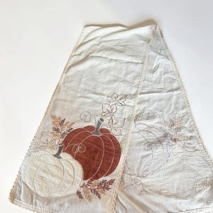 Peut inclure: Un chemin de table en tissu crème orné de motifs de citrouilles brodés. Un côté présente une citrouille blanche texturée et une citrouille couleur rouille avec du feuillage brun, tandis que l'autre côté montre un contour subtil de citrouilles.