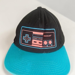 May include: A black baseball cap with a teal brim featuring a graphic print of a classic Nintendo game controller on the front panel. The controller design is outlined in light blue and filled with coral pink, including the text Nintendo.