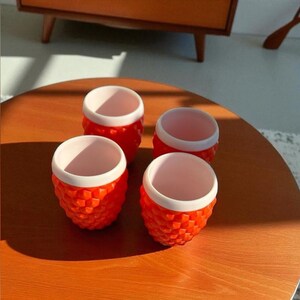 Peut inclure: Un ensemble de quatre petites tasses avec un extérieur orange vif présentant un motif géométrique texturé rappelant l'ananas et un bord intérieur blanc uni et lisse. Les tasses sont de forme arrondie avec un design uniforme.