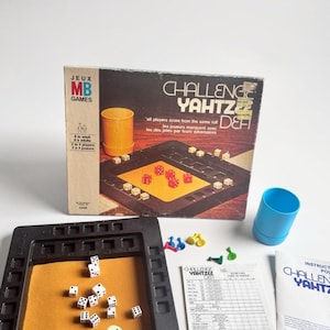 Peut inclure: Un jeu de société Challenge Yahtzee comprenant un plateau de jeu carré en plastique noir avec un centre en feutrine beige, des dés blancs à points noirs, un gobelet bleu, des petits pions en plastique colorés, une fiche de score et la boîte.